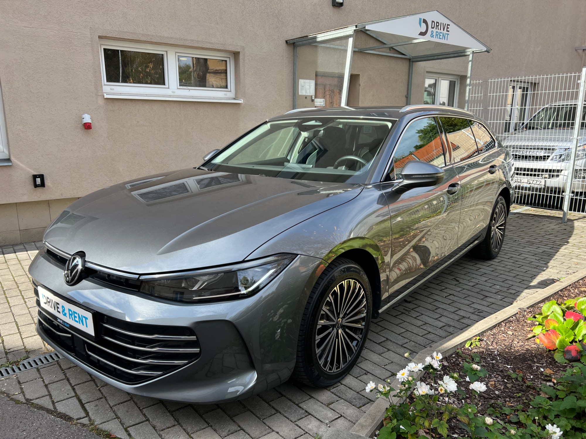 Šedý Volkswagen Passat Elegance zepředu a z boku s vícepaprskovými koly a střešními ližinami